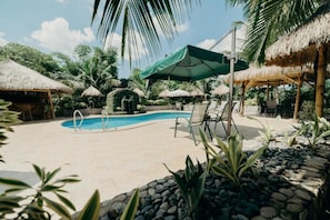 A pool area with lounge chairs and umbrellas, surrounded by tropical plants and thatched-roof structures. - La Casa Denika (Puerto Princesa)