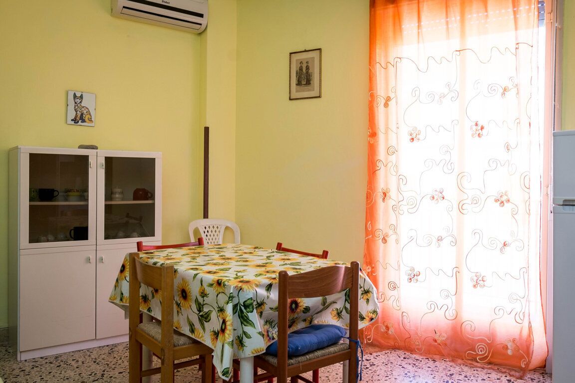 A dining area with a table covered by a sunflower tablecloth, a white cabinet with glass doors, and a chair with a blue cushion.