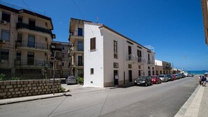 A coastal street with multi-story buildings, parked cars, and a clear blue sky.