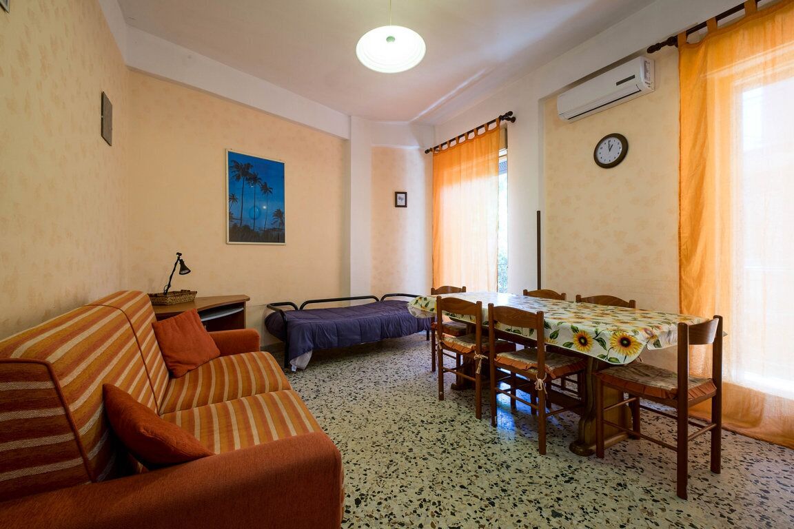 A cozy room with a striped sofa, a small bed, a dining table with chairs, a clock on the wall, and a window with curtains.