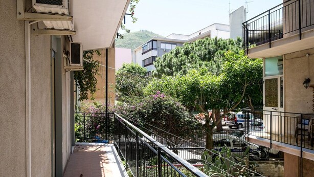 A narrow balcony with a metal railing, surrounded by greenery and a parked car.
