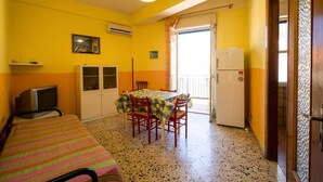 Una habitación compacta con cama, una mesa de comedor con sillas, un pequeño televisor, aire acondicionado y refrigerador.