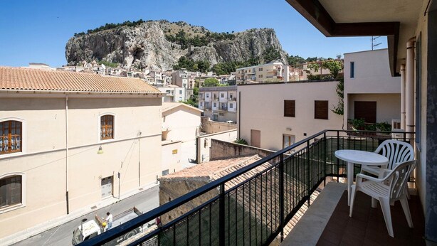 Un balcón con mesa y sillas, con vistas a un pueblo con edificios y una colina rocosa destacada al fondo.