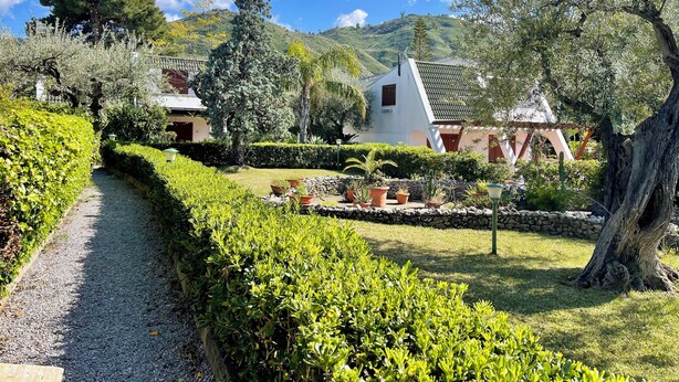Un sendero conduce a un edificio con techo inclinado, rodeado de setos cuidados y diversas plantas.