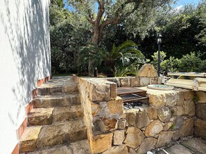 Un espacio al aire libre de piedra con una parrilla y asientos, rodeado de abundante vegetación y árboles.