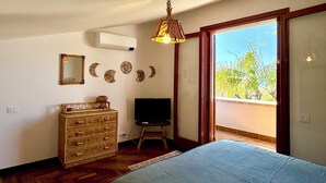 Un dormitorio con cama, cómoda, televisor, elementos decorativos en la pared y un ventanal con vistas a zonas verdes.