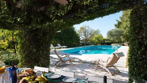 Un coin repas aménagé au bord de la piscine, avec une table dressée pour deux personnes, comprenant des assiettes, des verres et un panier de fruits.