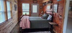 A wooden cabin bedroom with a bed, a nightstand, a lamp, and a window with blinds.