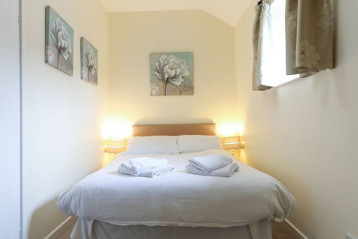 A neatly made bed with white linens and towels, two wall-mounted paintings of flowers, and a bedside lamp.