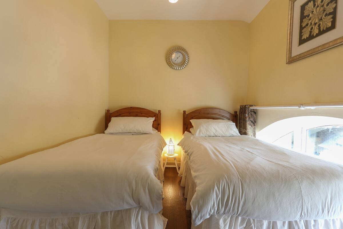 A room with two single beds, a small table with a lamp, a wall clock, and a framed picture.