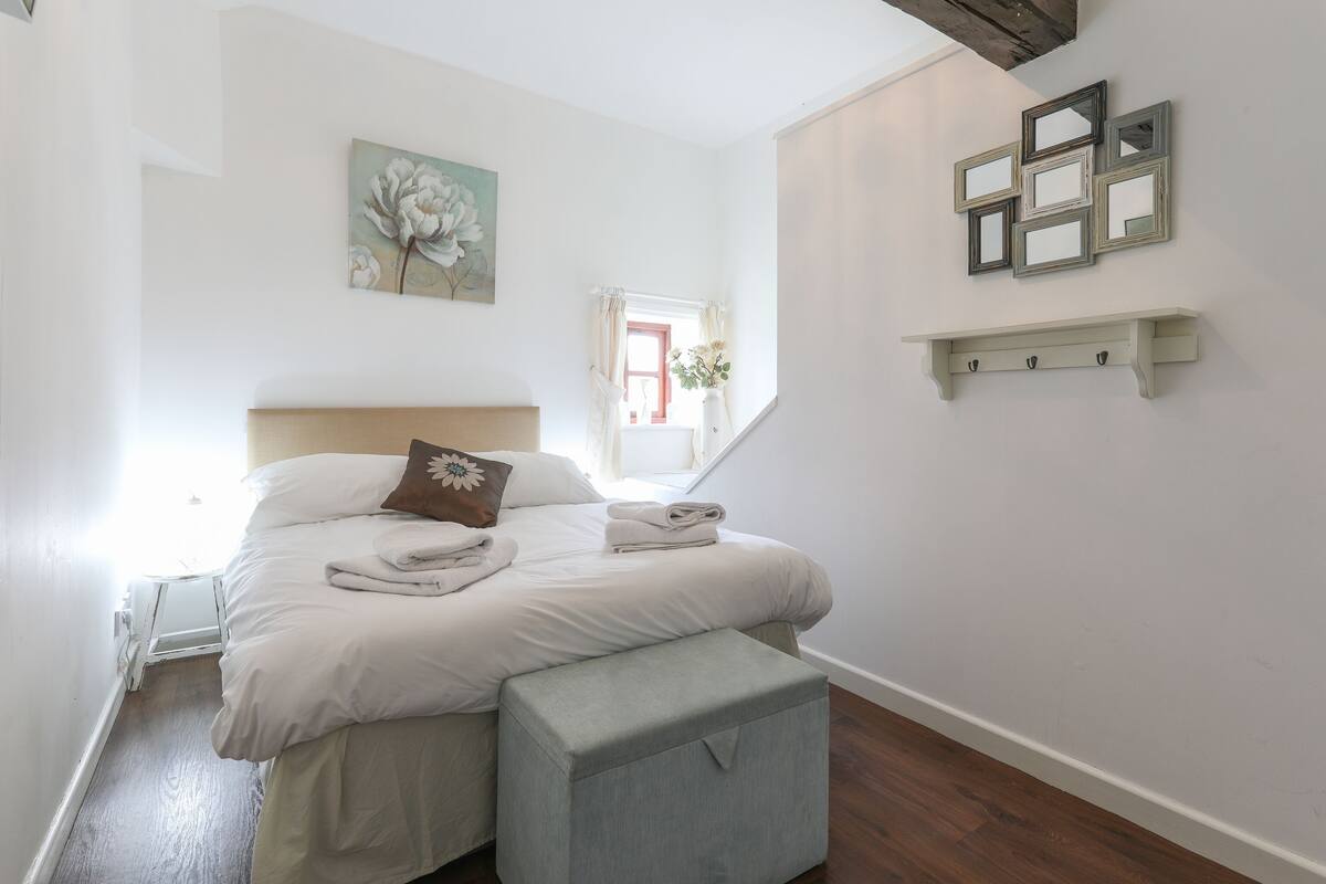 A neatly made bed with a pillow, a bedside table, a small bench, and a wall-mounted shelf with mirrors.
