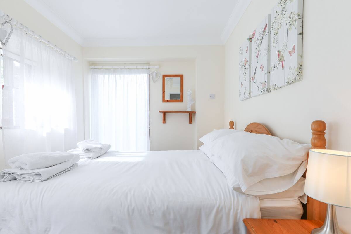 A hotel room with a neatly made bed, a bedside table with a lamp, a small wooden shelf, and a large window with sheer curtains.