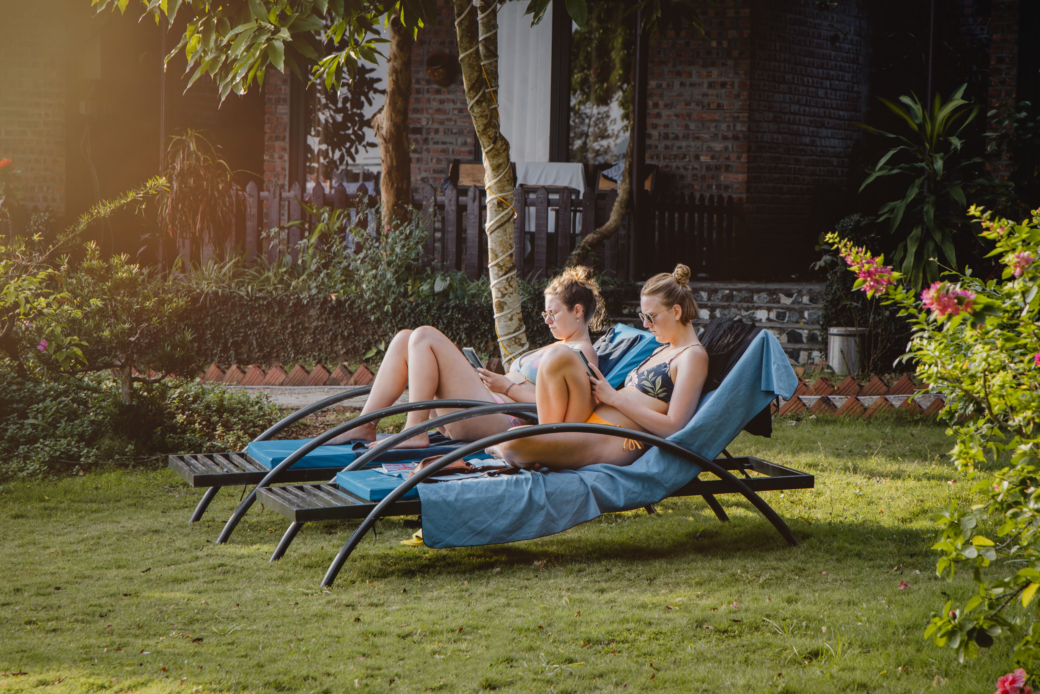 Two people relaxing on a hammock chair in a garden with a brick wall and greenery in the background.
