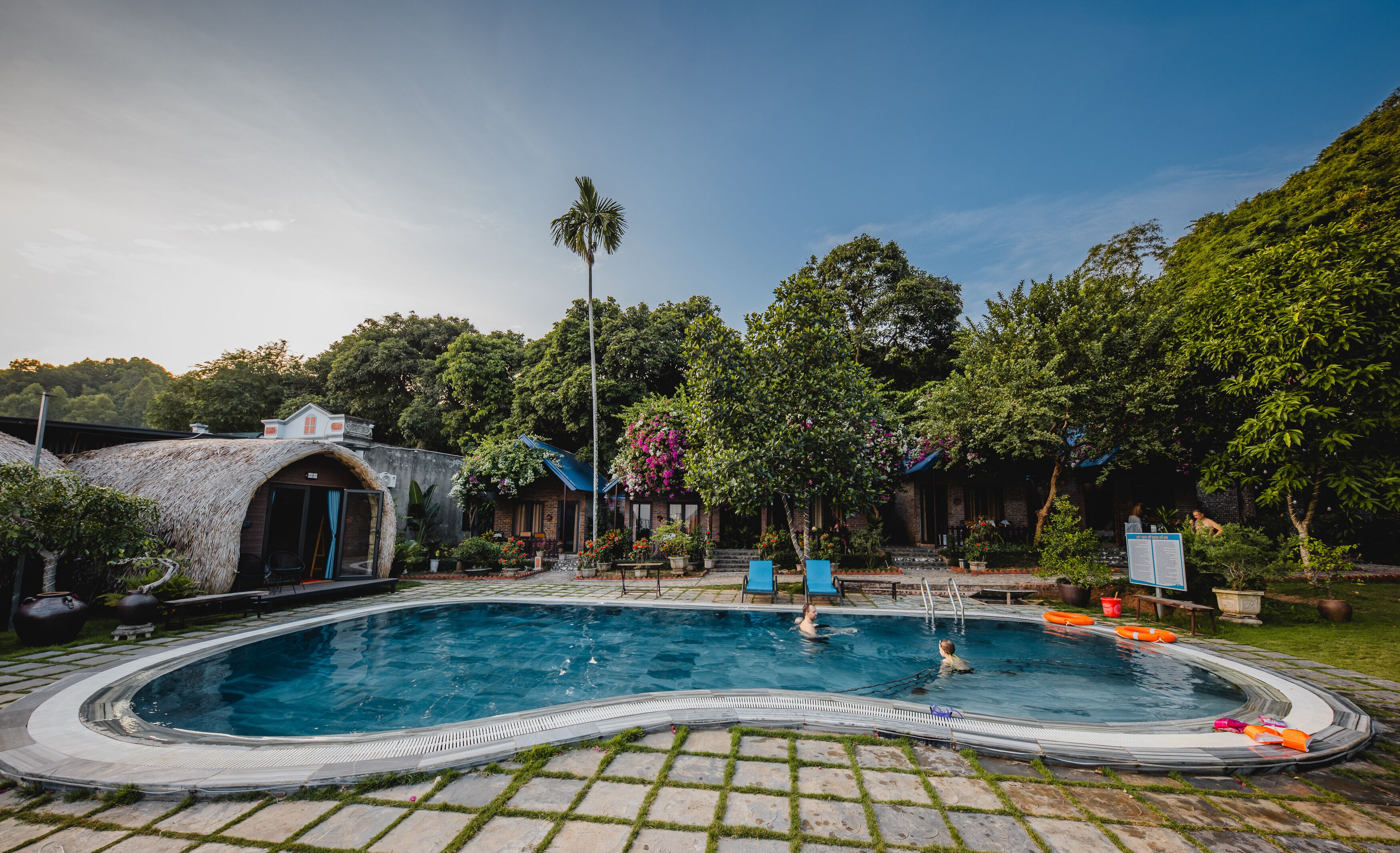 A swimming pool with a hot tub, surrounded by lush greenery and a brick building.