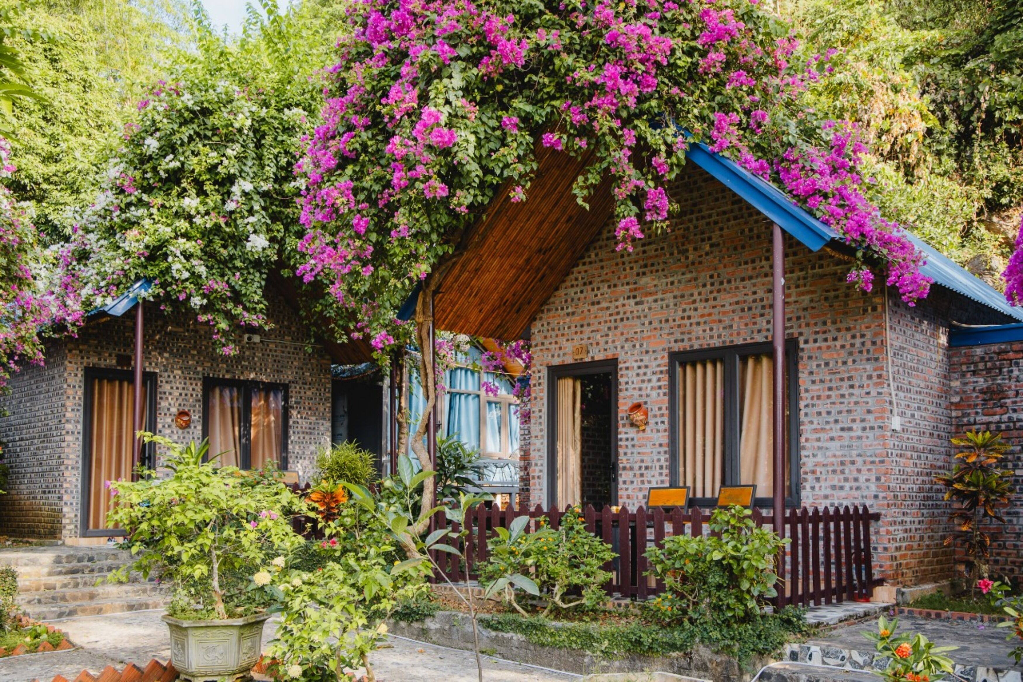 A rustic brick house with a wooden porch, surrounded by lush greenery and flowering plants.