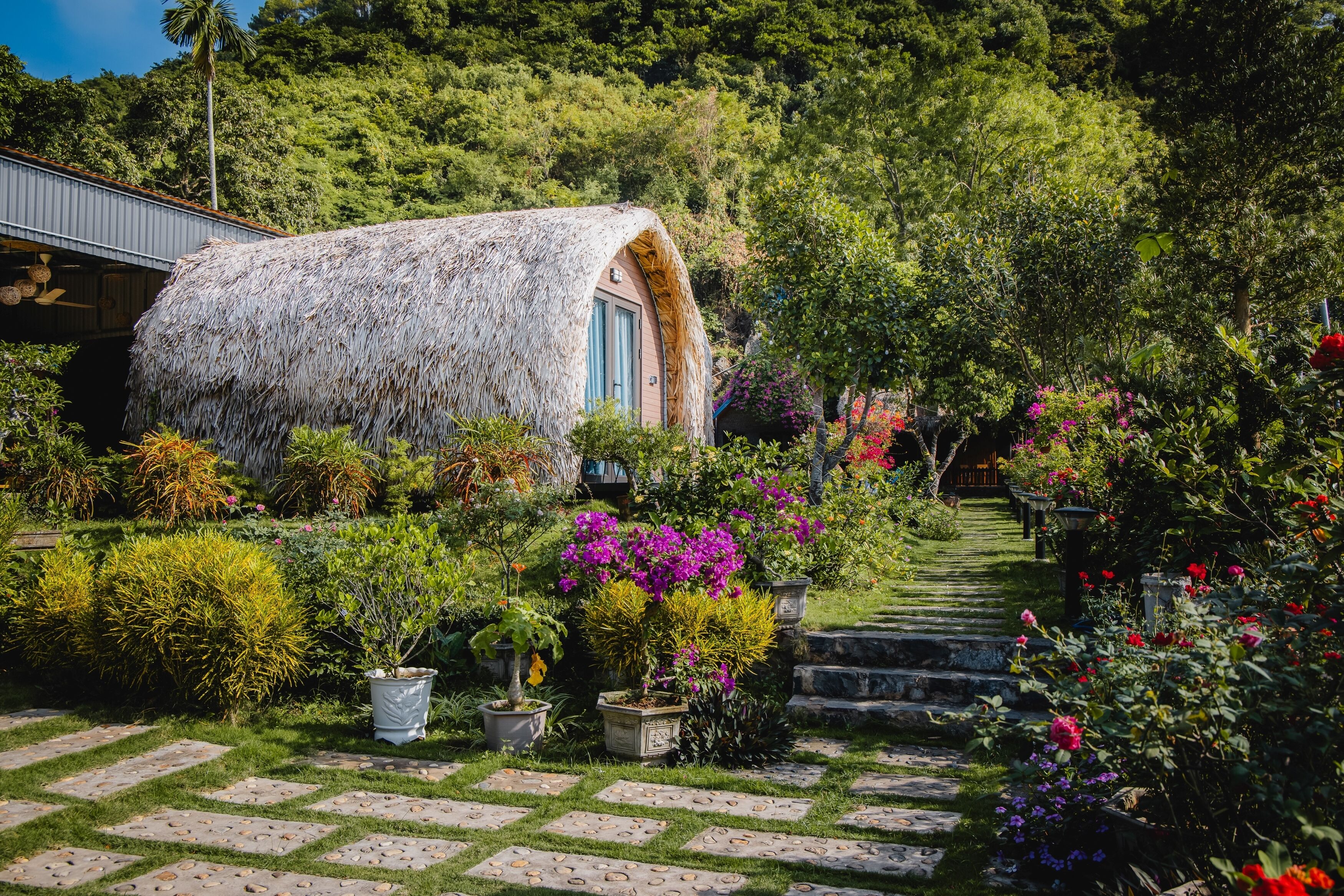 A thatched-roof structure with a door, surrounded by a garden with various plants and a stone pathway.