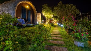 A thatched-roof structure with a pathway leading to it, surrounded by lush greenery and flowering plants. - Green Homestay (Hai Phong)