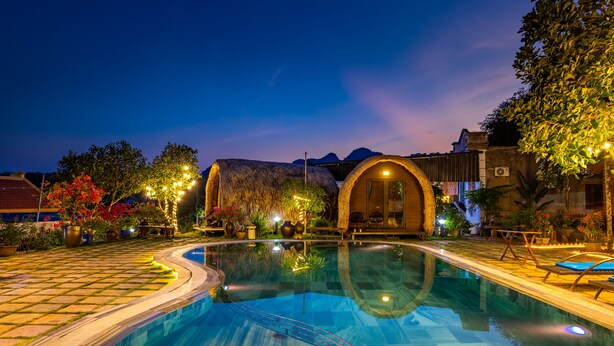 A swimming pool with a curved edge, surrounded by a paved area with a thatched-roof structure and a small garden with potted plants. - Green Homestay (Hai Phong)