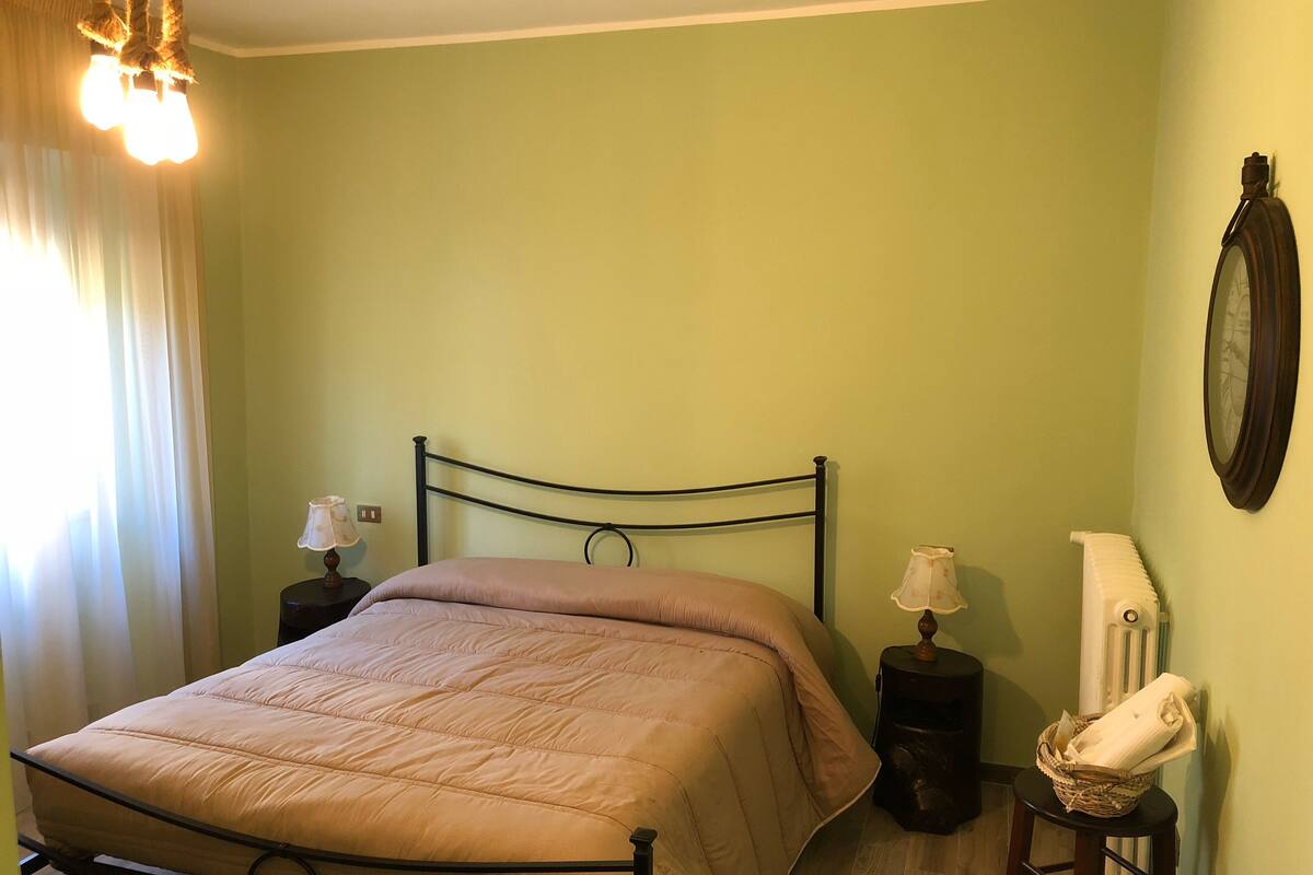 Una cama con cabecera, dos mesitas de noche con lámparas, un radiador y un reloj de pared.