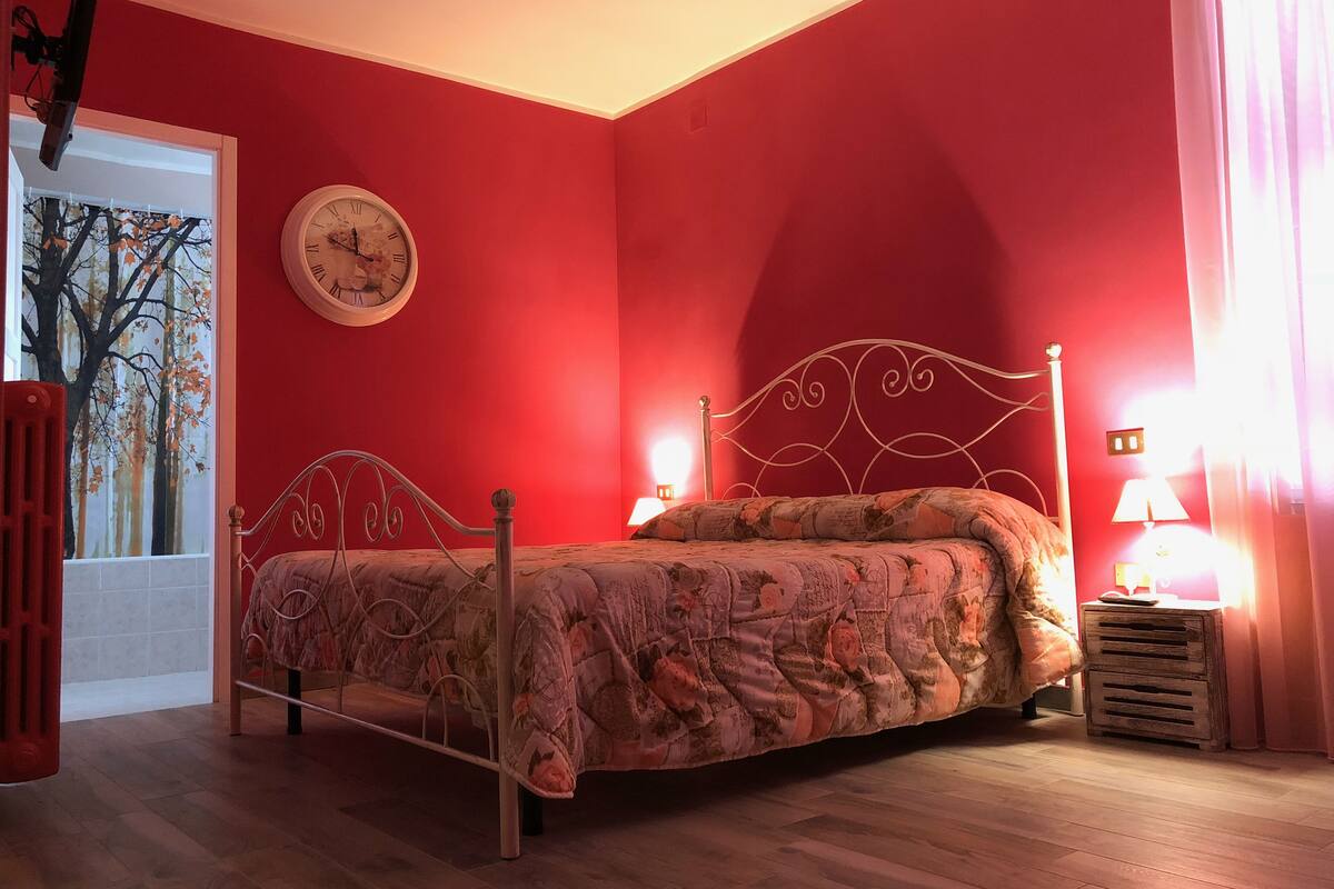 Un dormitorio con pared roja, cama con colcha estampada, un reloj de pared redondo y una ventana con un cuadro de un árbol.