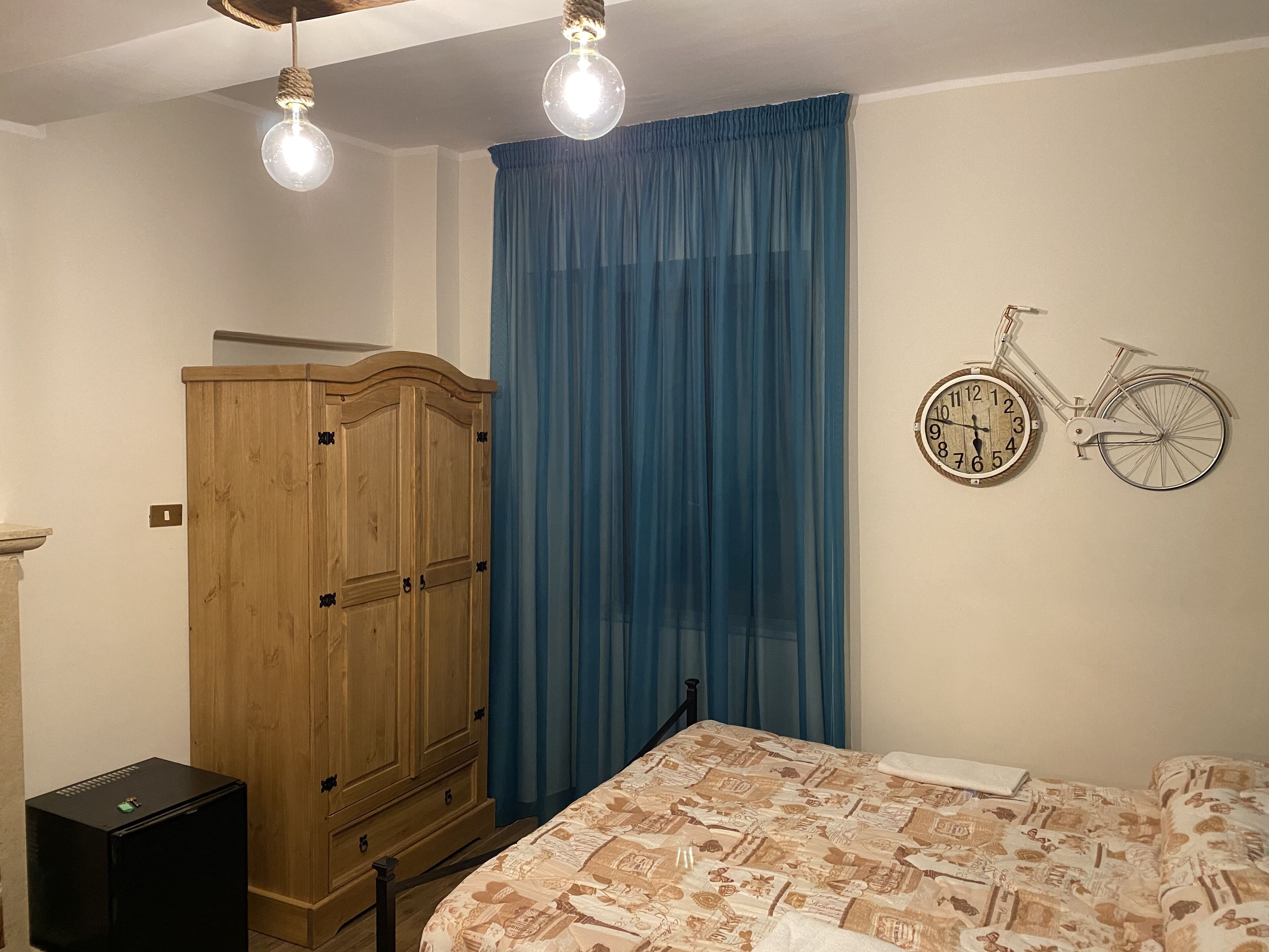 A bedroom with a bed, a wooden wardrobe, a black speaker, and a wall clock shaped like a bicycle.