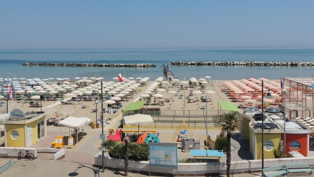 Une vue sur la plage, avec de nombreux parasols et des chaises longues, une digue rocheuse et un ciel bleu dégagé.