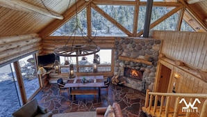 L’intérieur d’une cabane en rondins, avec une cheminée en pierre, des meubles en bois et une grande fenêtre donnant sur un paysage enneigé.