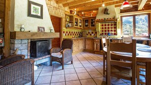A rustic cabin interior with a stone fireplace, wicker furniture, wooden beams, and a kitchen area with a checkered backsplash.