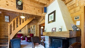 A cozy log cabin interior with a fireplace, wicker furniture, a wooden staircase, and a clock on the wall.
