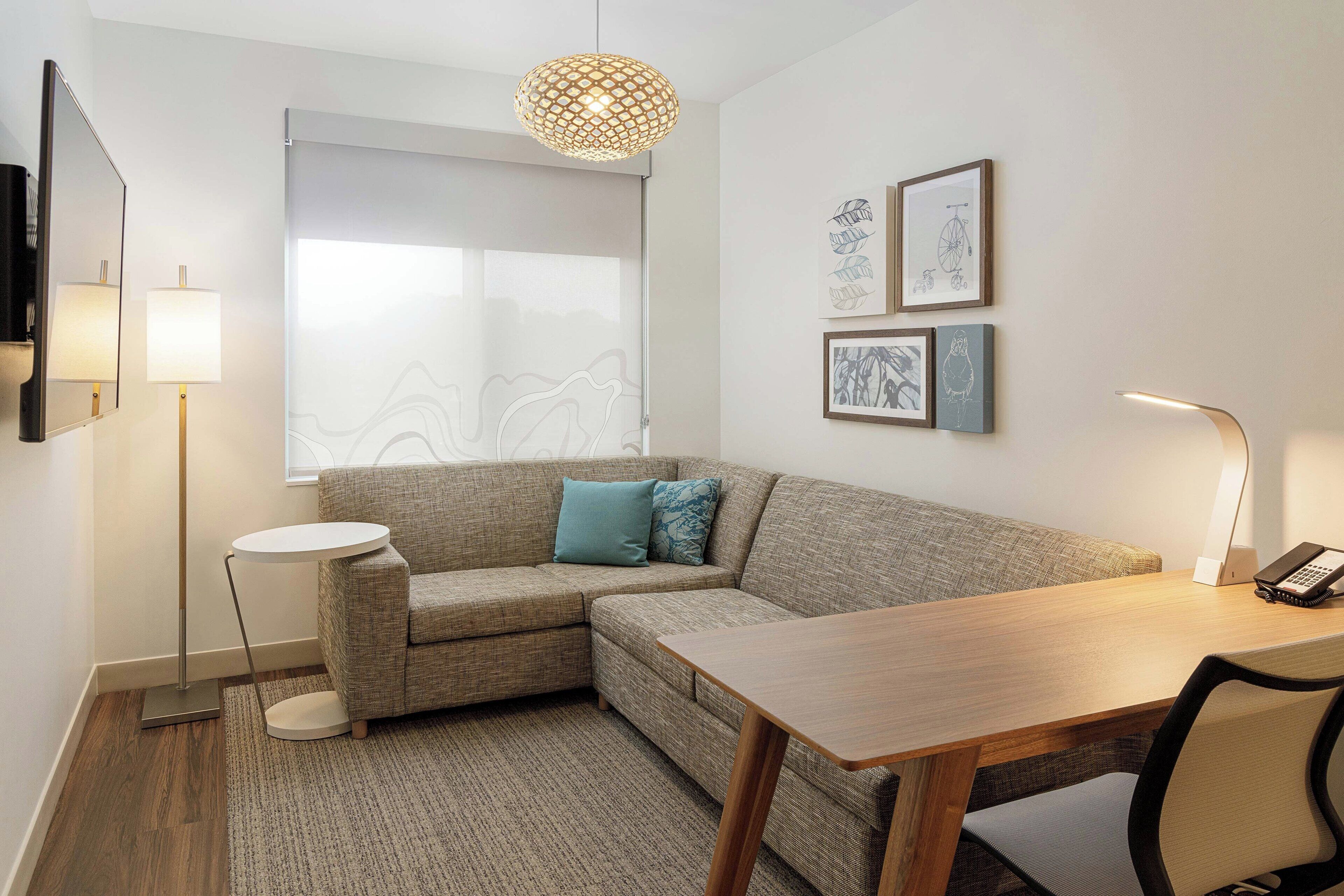 Una habitación de hotel moderna con un sofá modular, una mesita redonda, un escritorio con una silla, una lámpara de pie y obras de arte en la pared.