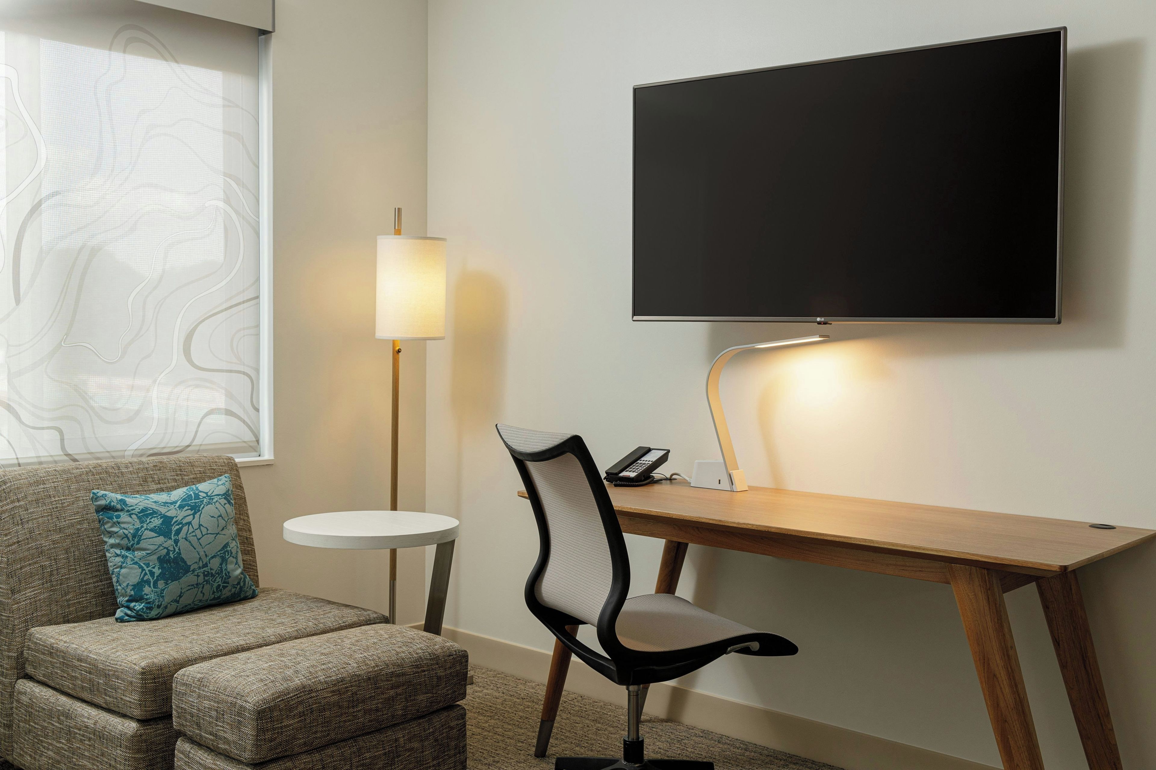 Een hotelkamer met een wandtelevisie, een bureau met stoel, een klein rond tafeltje en een bank met een gestreept kussen.