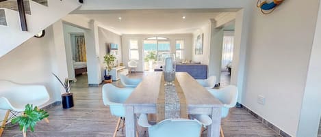 Un vaste espace de salle à manger avec une longue table en bois et des chaises modernes, qui donne sur une cuisine et un salon agrémenté d’un escalier.