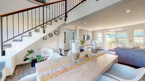 A modern living room with a staircase, a dining area with a long table, and a view of the outdoors. - Caribbean Estate Deluxe (Port Edward)