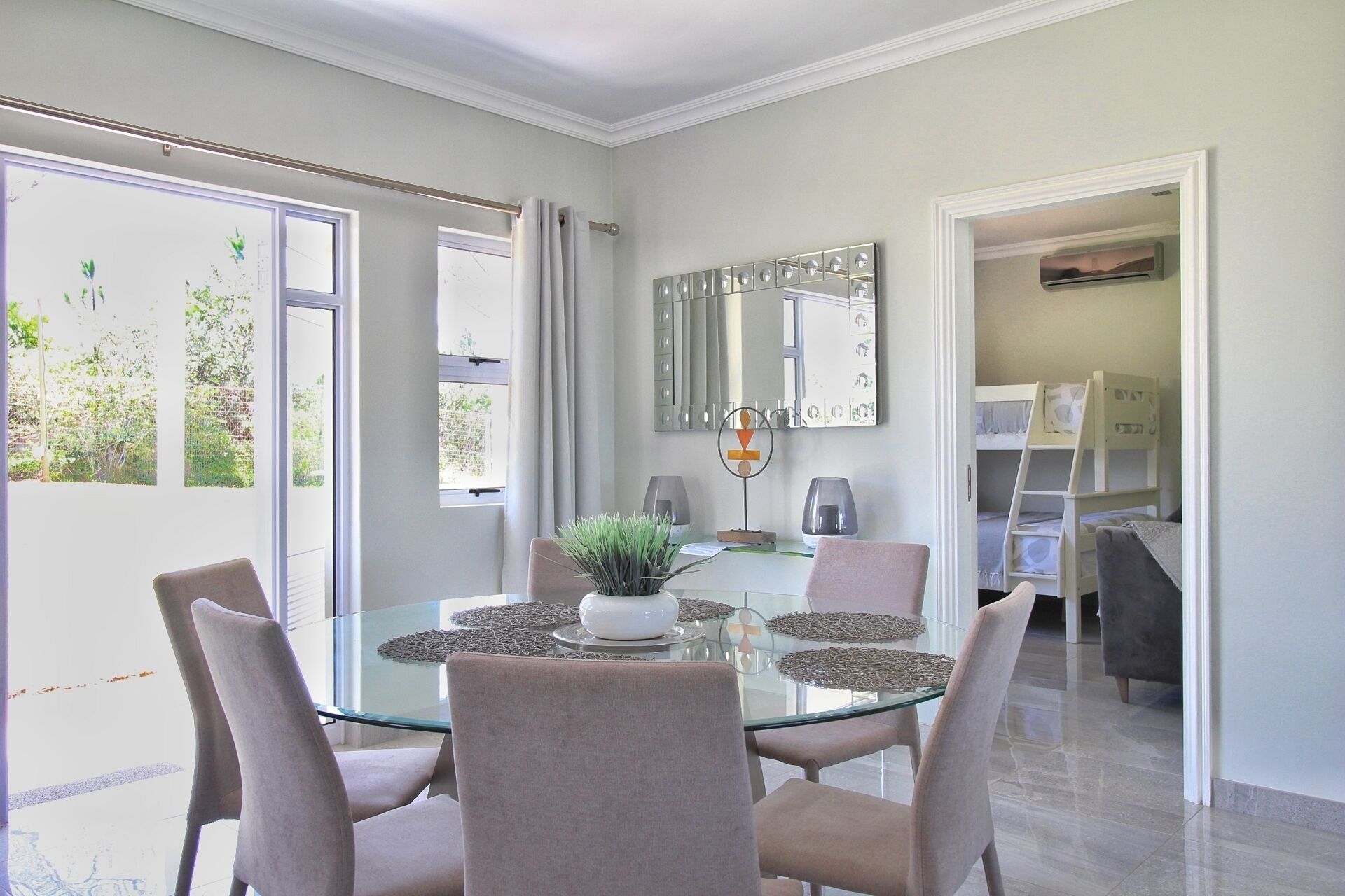A dining area with a glass table surrounded by chairs, a potted plant, and a decorative mirror.