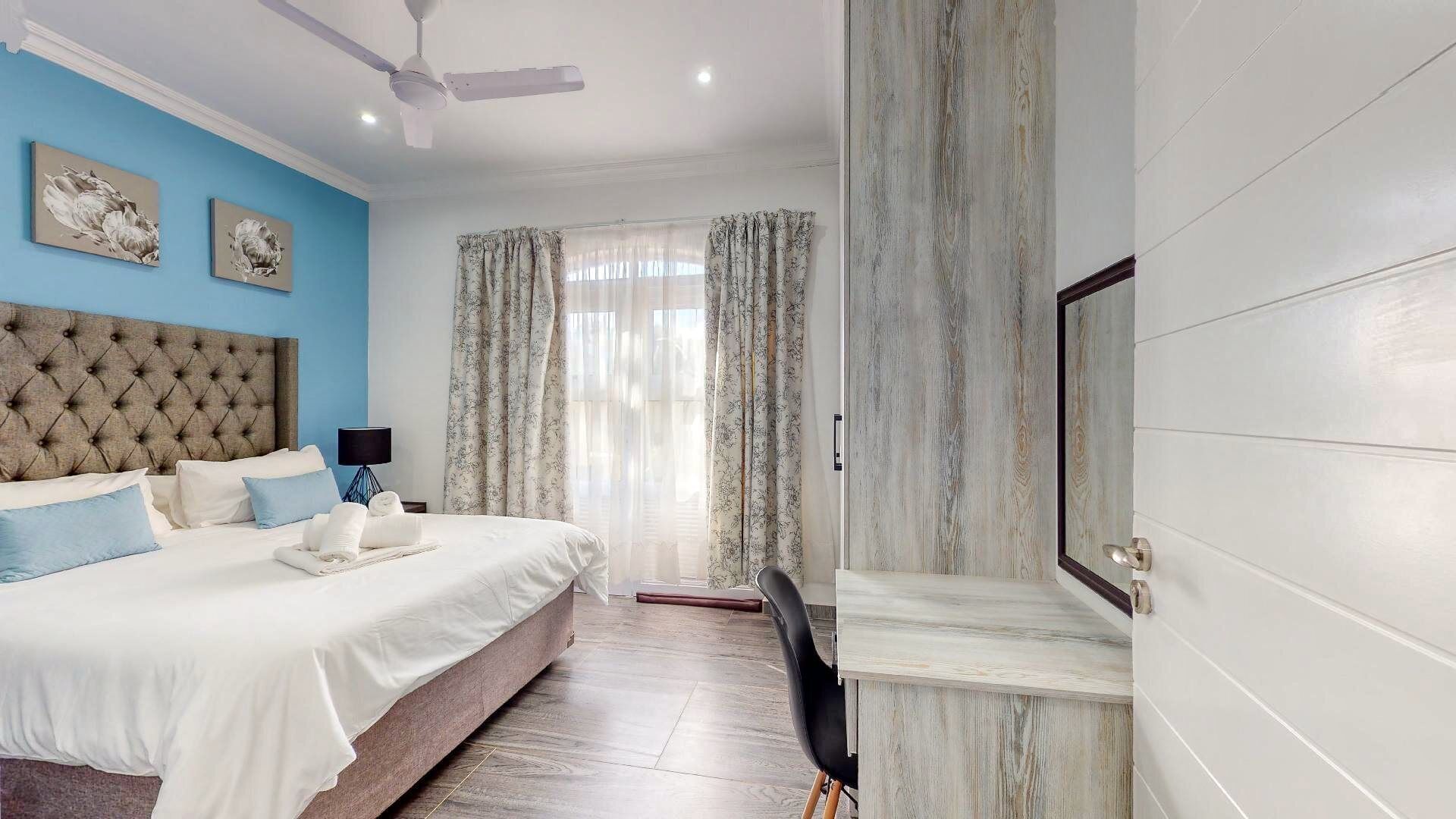 Une chambre à coucher avec une tête de lit capitonnée, un lit avec une literie blanche et bleue, un ventilateur de plafond et un téléviseur fixé au mur.