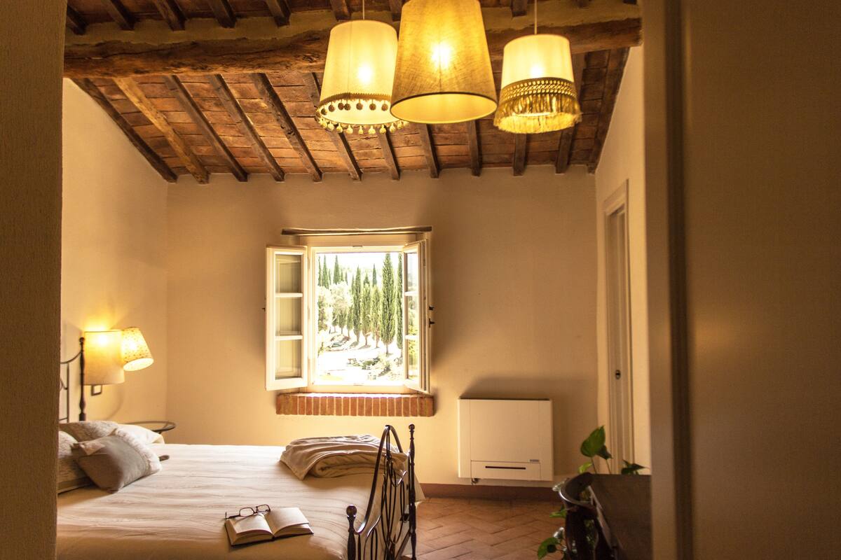 Ein Schlafzimmer mit Bett, Nachttischlampen, einem Fenster mit Blick auf Bäume und einer Heizung.