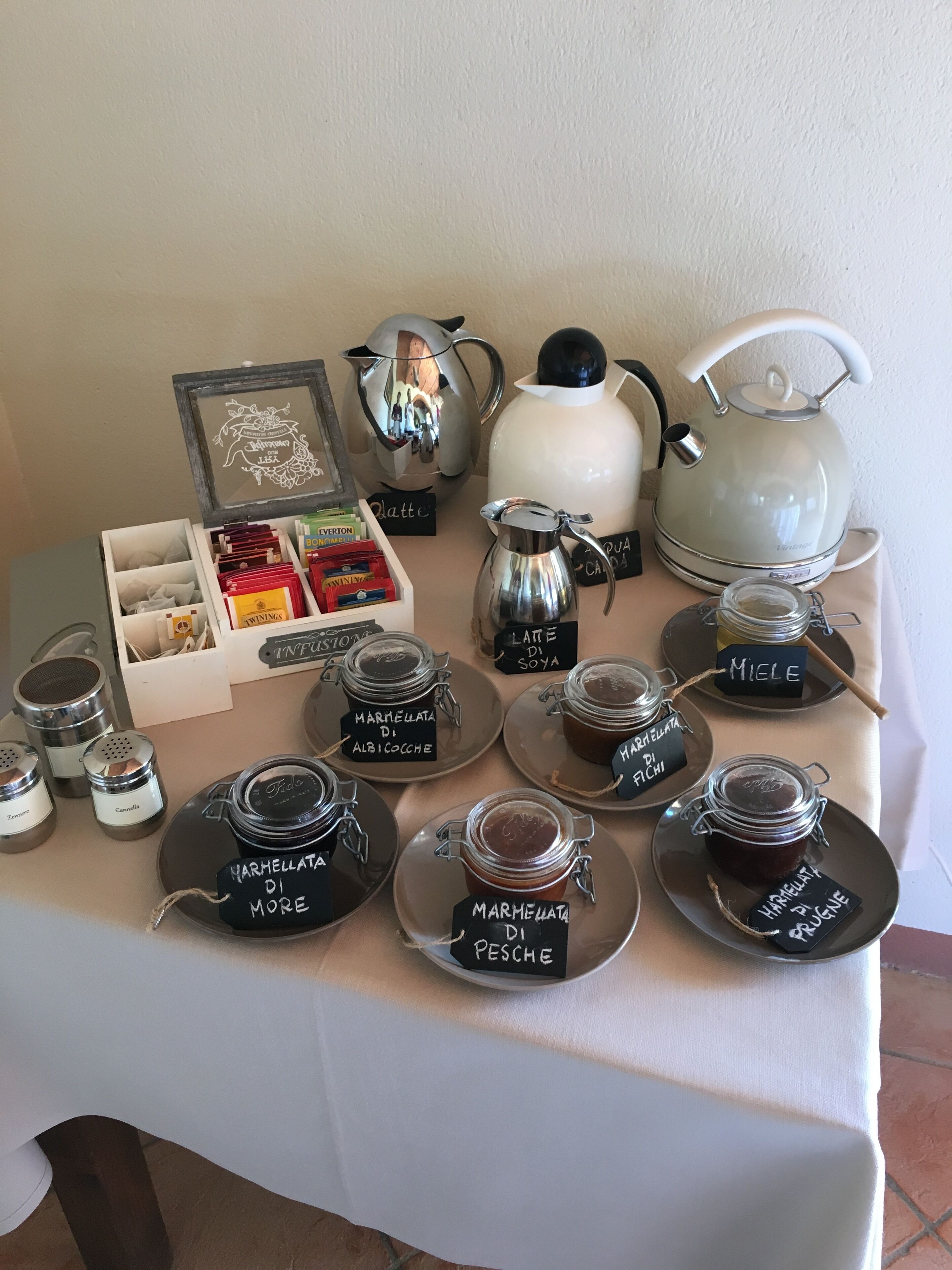 Una mesa con diferentes opciones de té y café, incluyendo una tetera blanca, un hervidor plateado y varios frascos de vidrio etiquetados con distintos tipos de mermelada.