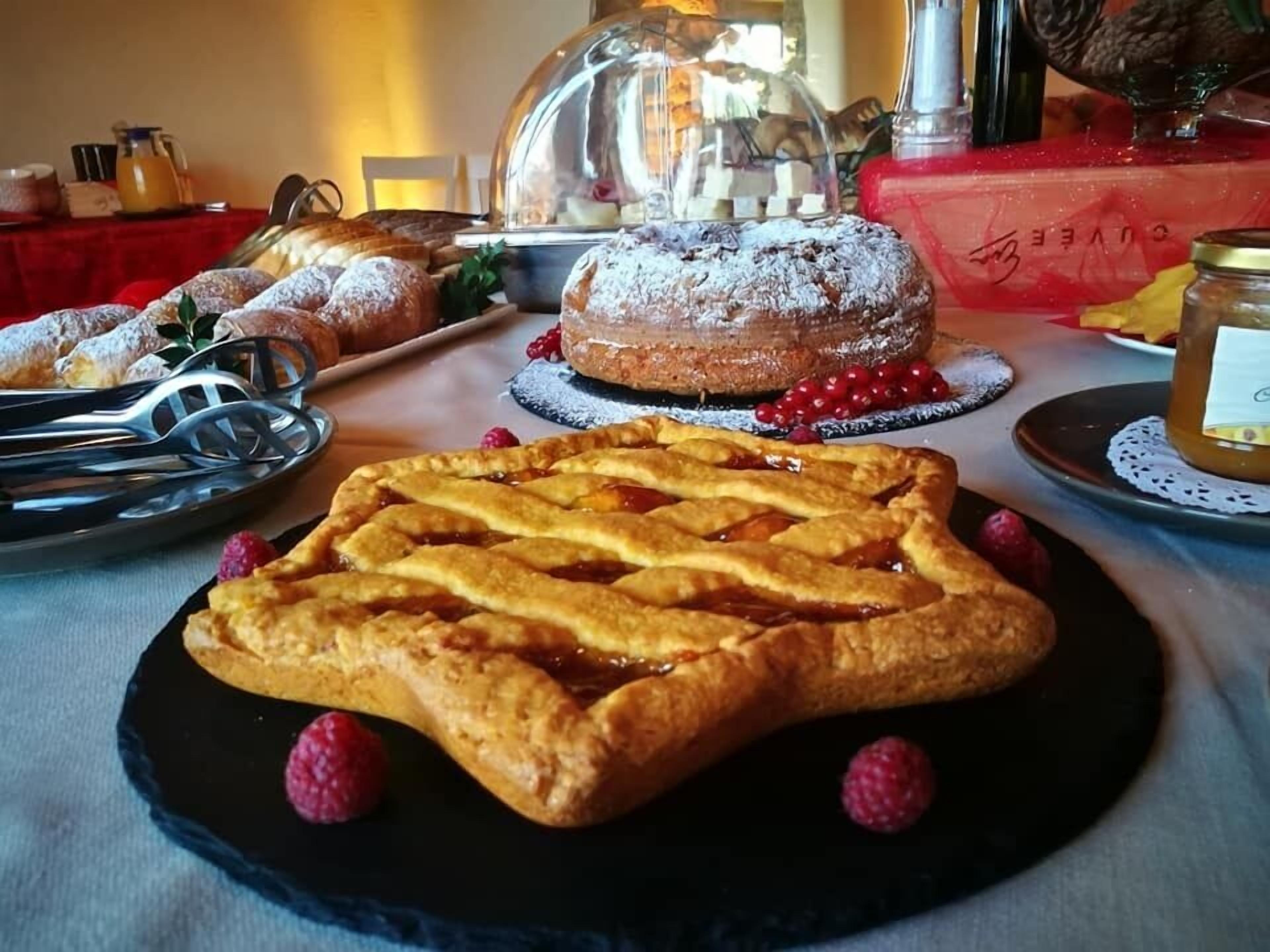 A table with various pastries, including a pie with a lattice crust and raspberries, a covered pastry case, and a jar of jam.