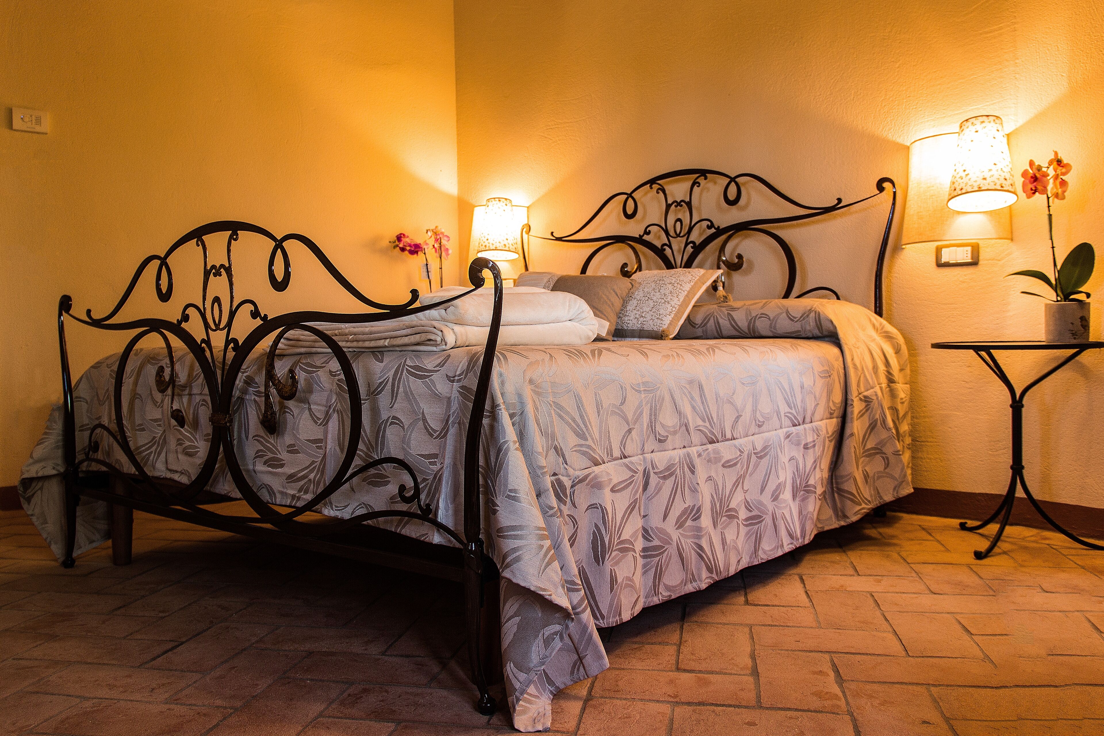 A neatly made bed with a floral-patterned bedspread, a metal headboard, and two bedside lamps on a nightstand with a potted plant.