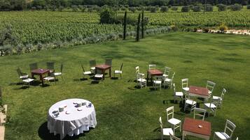 Un área de comedor al aire libre con varias mesas y sillas, rodeada de un césped verde exuberante y viñedos al fondo.