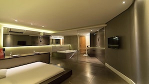 Una habitación de hotel moderna con una cama grande, una zona de estar con una mesita y sillas, un televisor montado en la pared y un baño separado.