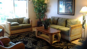 Una sala de estar con dos sofás verdes, una mesa de centro, una planta grande en maceta, un cuadro en la pared y una lámpara de pie.