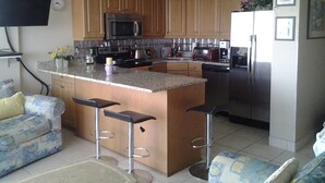 Una cocina con armarios de madera, una isla central con taburetes, microondas, refrigerador y televisor de pantalla plana montado en la pared.