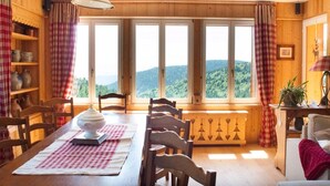 A wooden dining room with a table set for four, large windows with a view of a forested landscape, and a shelf with pottery.
