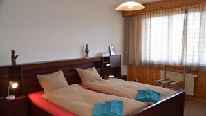 A double bed with two pillows and blue towels, wooden headboard, bedside tables with lamps, a small cabinet, and a window with curtains. - Nice flat to rent in les Collons (Thyon-Les Collons)