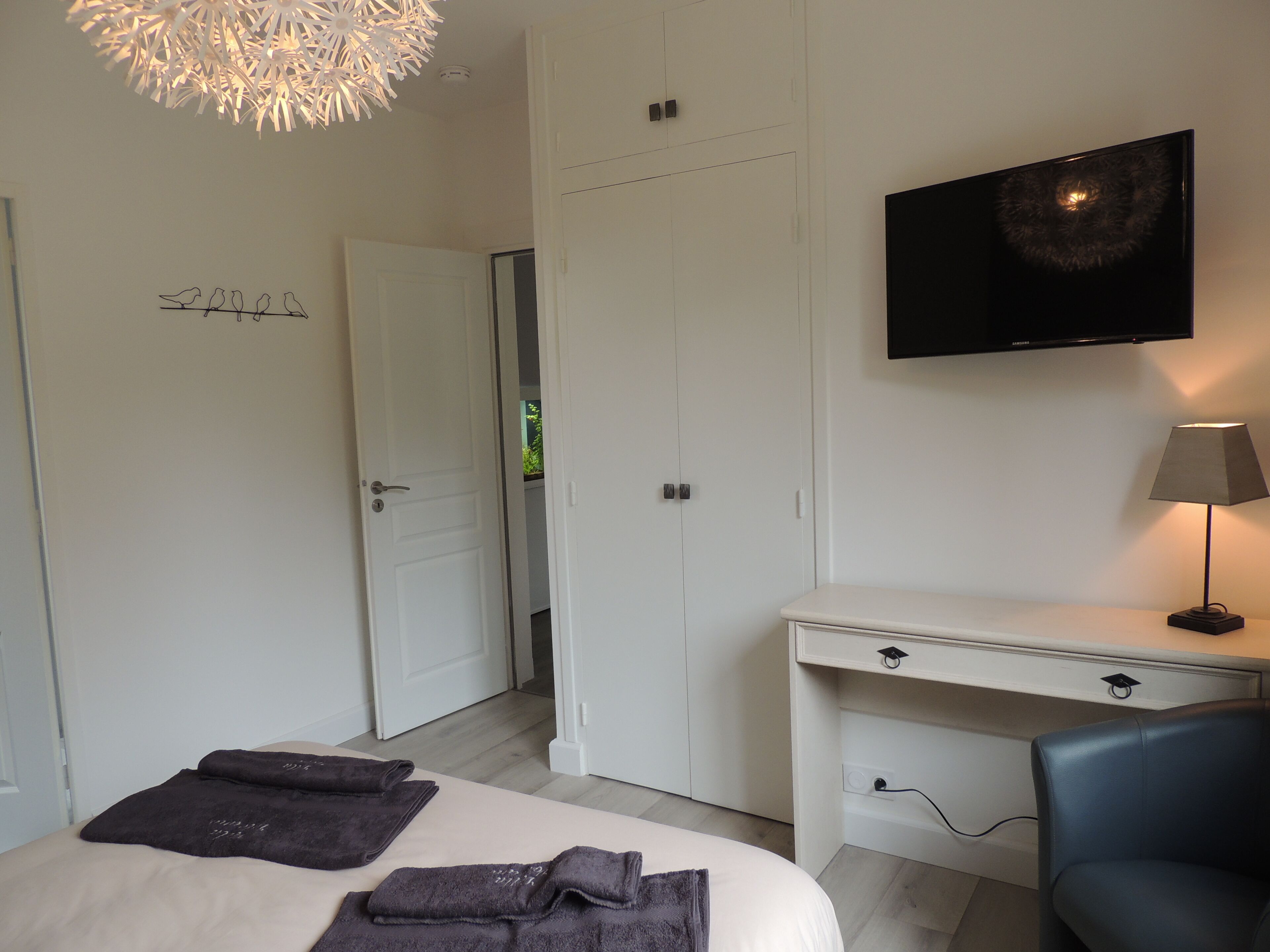 A bedroom with a bed, a desk, a chair, a TV, and a door leading to another room.