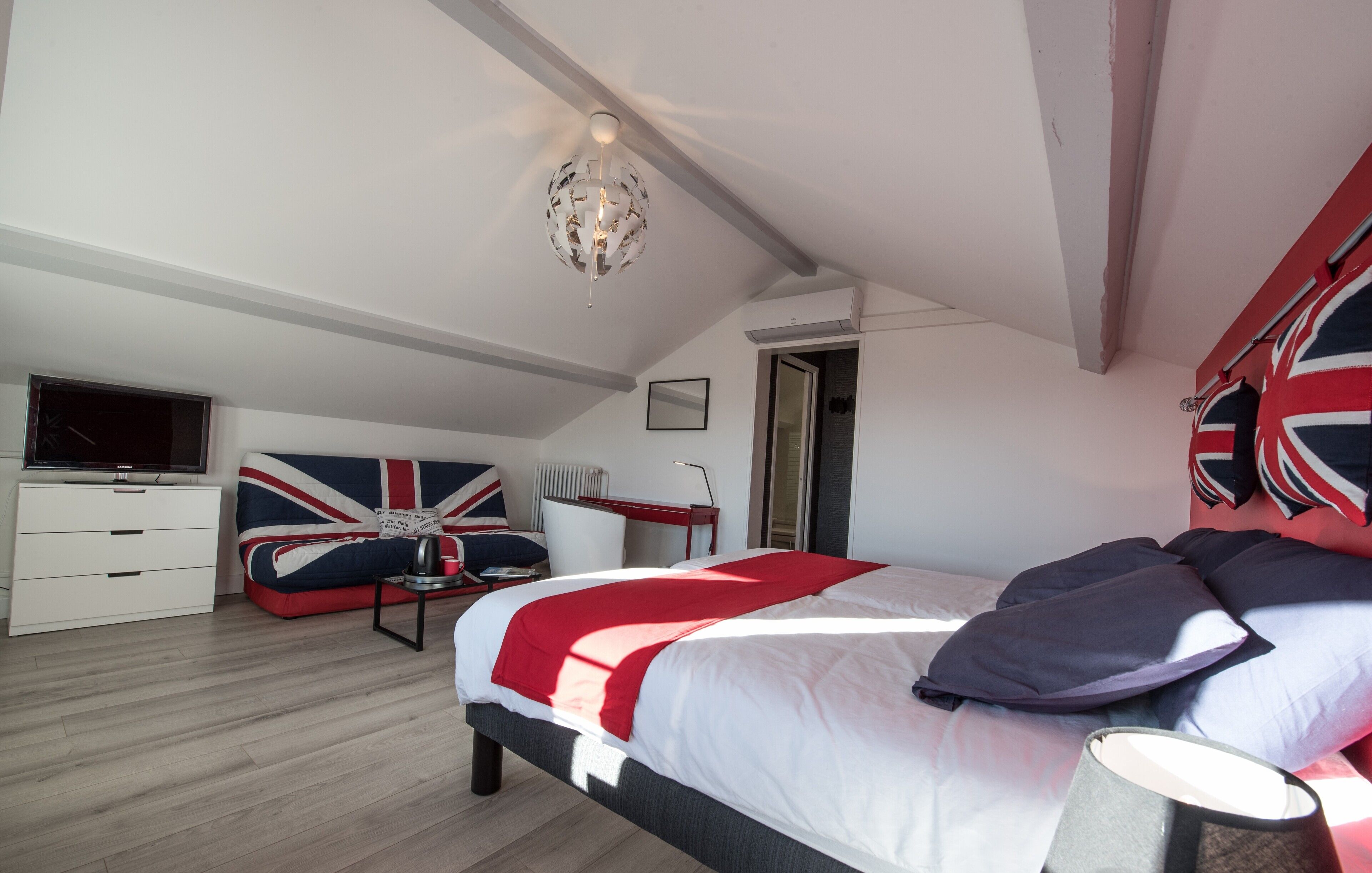 Un dormitorio con cama, televisor, escritorio y silla. Hay una ventana y una puerta.