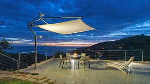 Een terras met een tafel, stoelen, een zonnescherm en uitzicht op zee in de schemering.