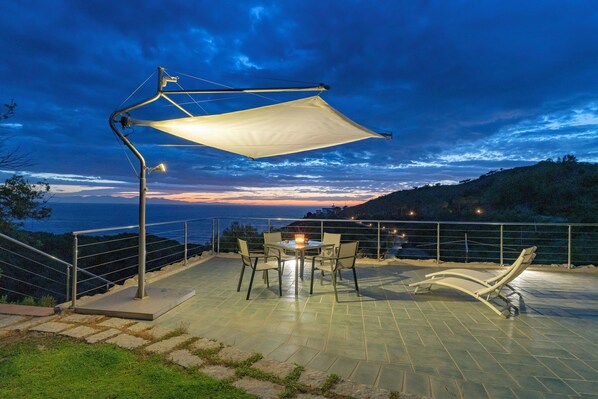 A terrace with a table and chairs, a sunshade, and a view of the sea at dusk. - Breathtaking sunset (marciana)