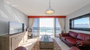 A modern living room with a large TV, a wooden coffee table, a red leather sofa, and a view of the ocean. - Luxury Unit with iconic breathtaking  views of Table Mountain & Robben island. (Bloubergstrand)